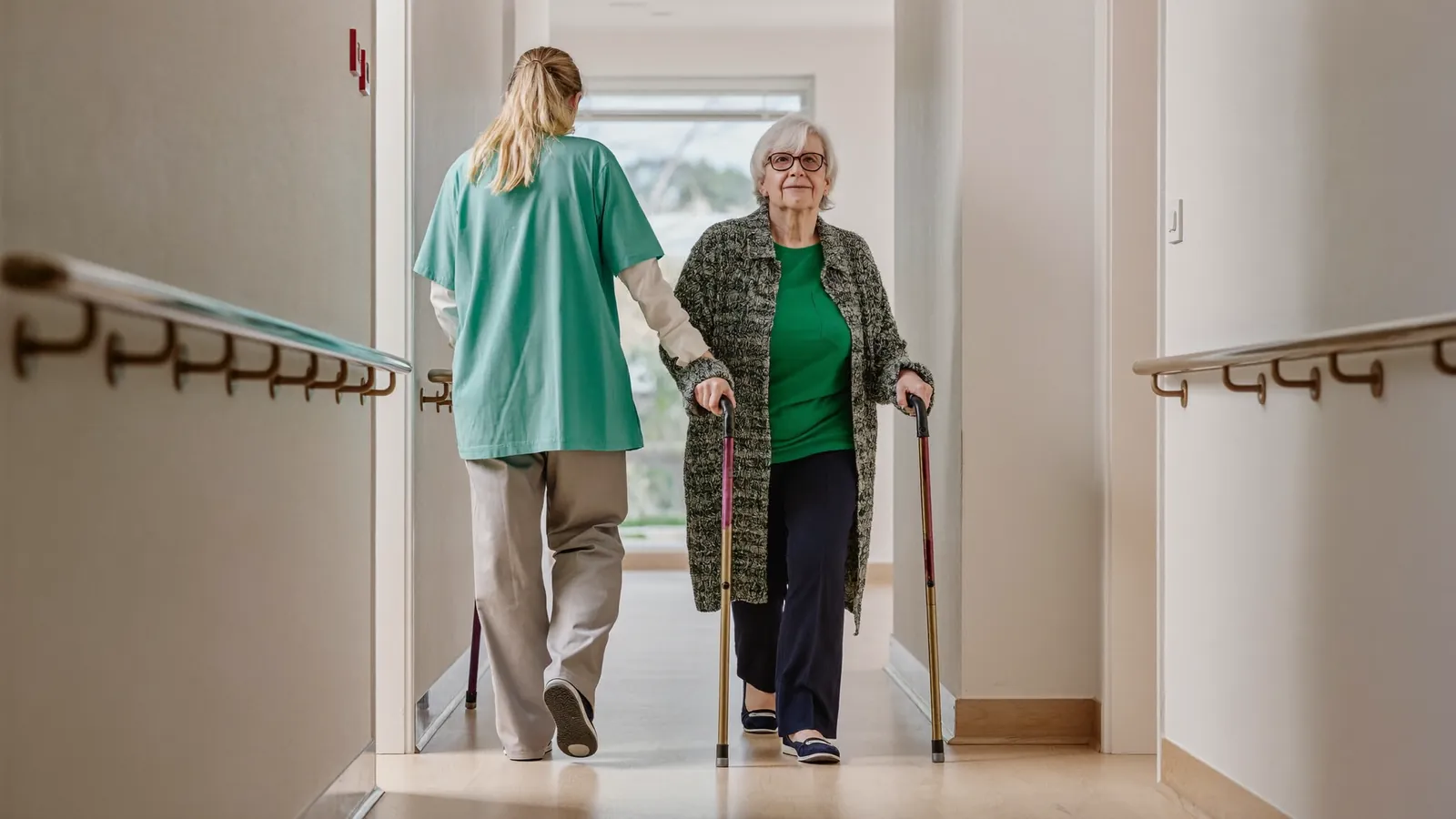 A caregiver walking beside an older woman with dementia down a bright hallway with handrails