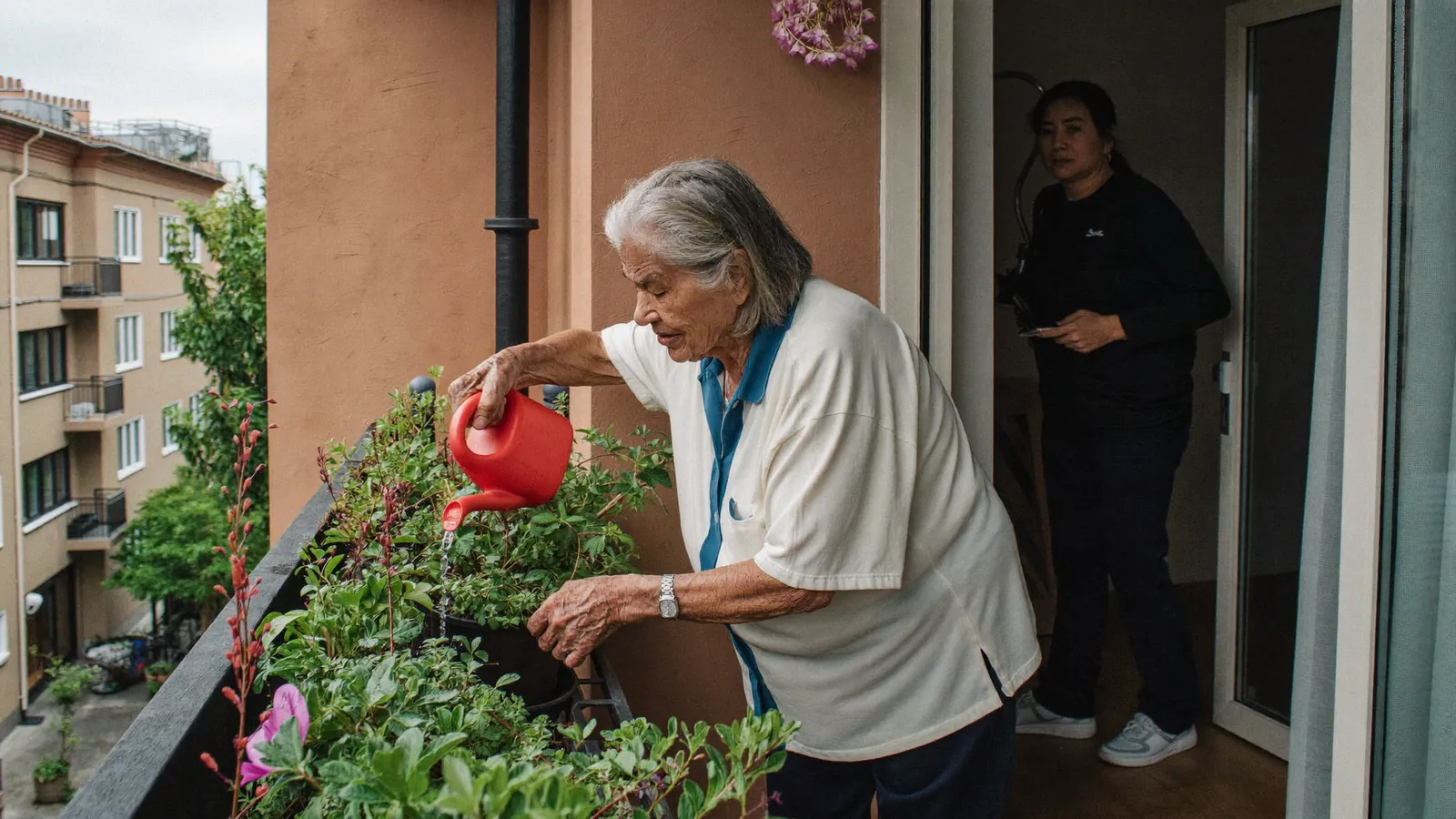 An older woman watering balcony plants while a caregiver visits at the door