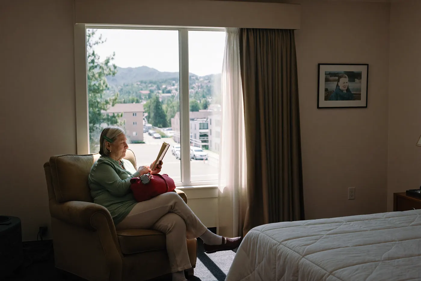 An older woman reads by the window of a short stay guest suite with an overnight bag packed